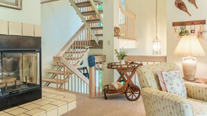 A cozy living room with a fireplace, a wooden staircase, a wicker chair, and a lamp.