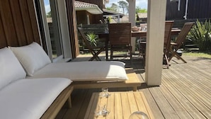 Een houten terras met een comfortabele zithoek en een tafel met wijnglazen en een bloevas.