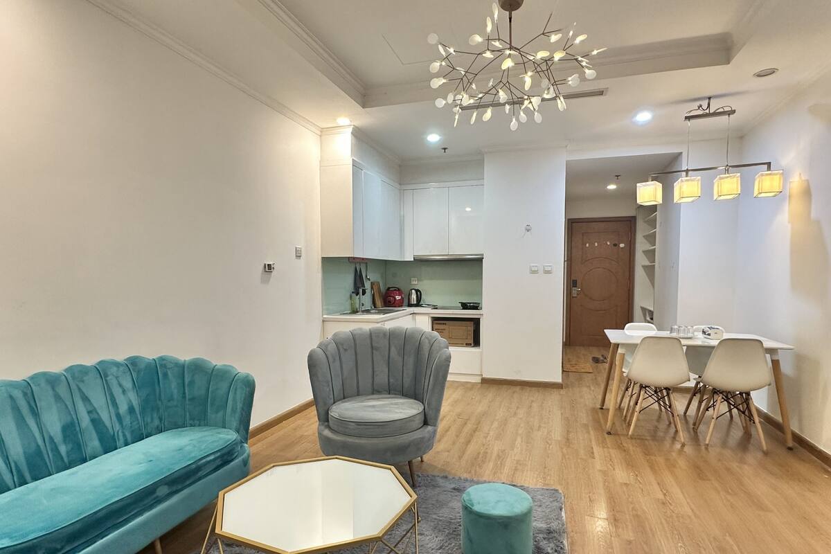 Un salon moderne avec un canapé turquoise, un fauteuil gris et une table basse en verre. On aperçoit une cuisine à l’arrière-plan, avec des meubles blancs, et un coin repas comprenant une table et des chaises.