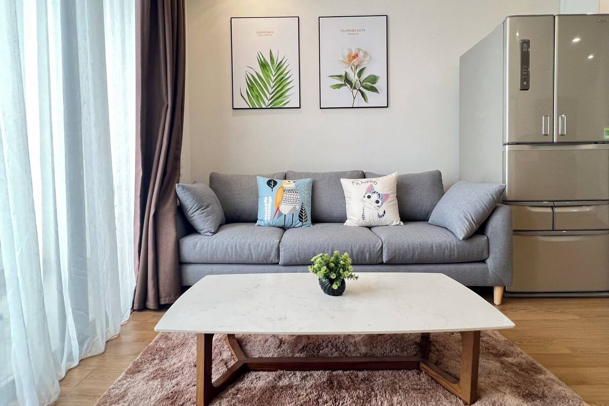 Un salon moderne avec un canapé gris, deux coussins, une table basse blanche et une petite plante. Deux reproductions botaniques encadrées ornent le mur.