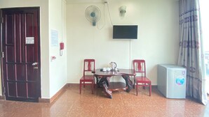 Una habitación con una mesa de comedor, dos sillas, un televisor montado en la pared, un ventilador, un refrigerador y una puerta con un cartel.