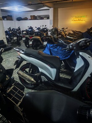 Un estacionamiento con varias motocicletas y scooters, un letrero de neón que indica un hotel, y un piso de baldosas.