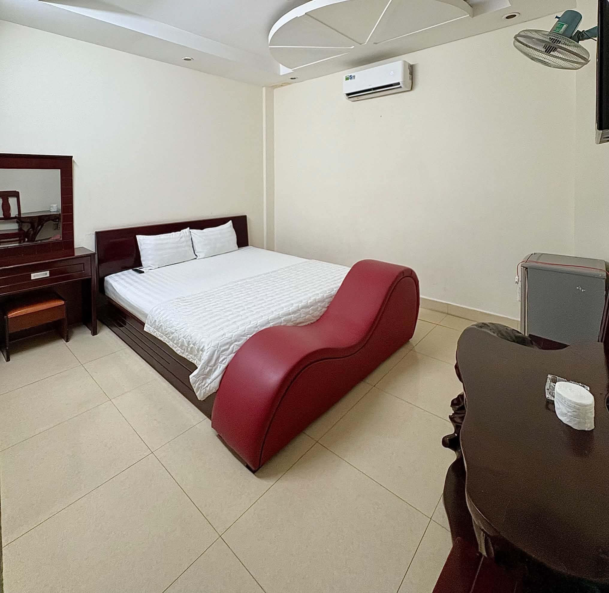 A hotel room with a bed, a desk, a chair, a mirror, an air conditioning unit, and a small cabinet.