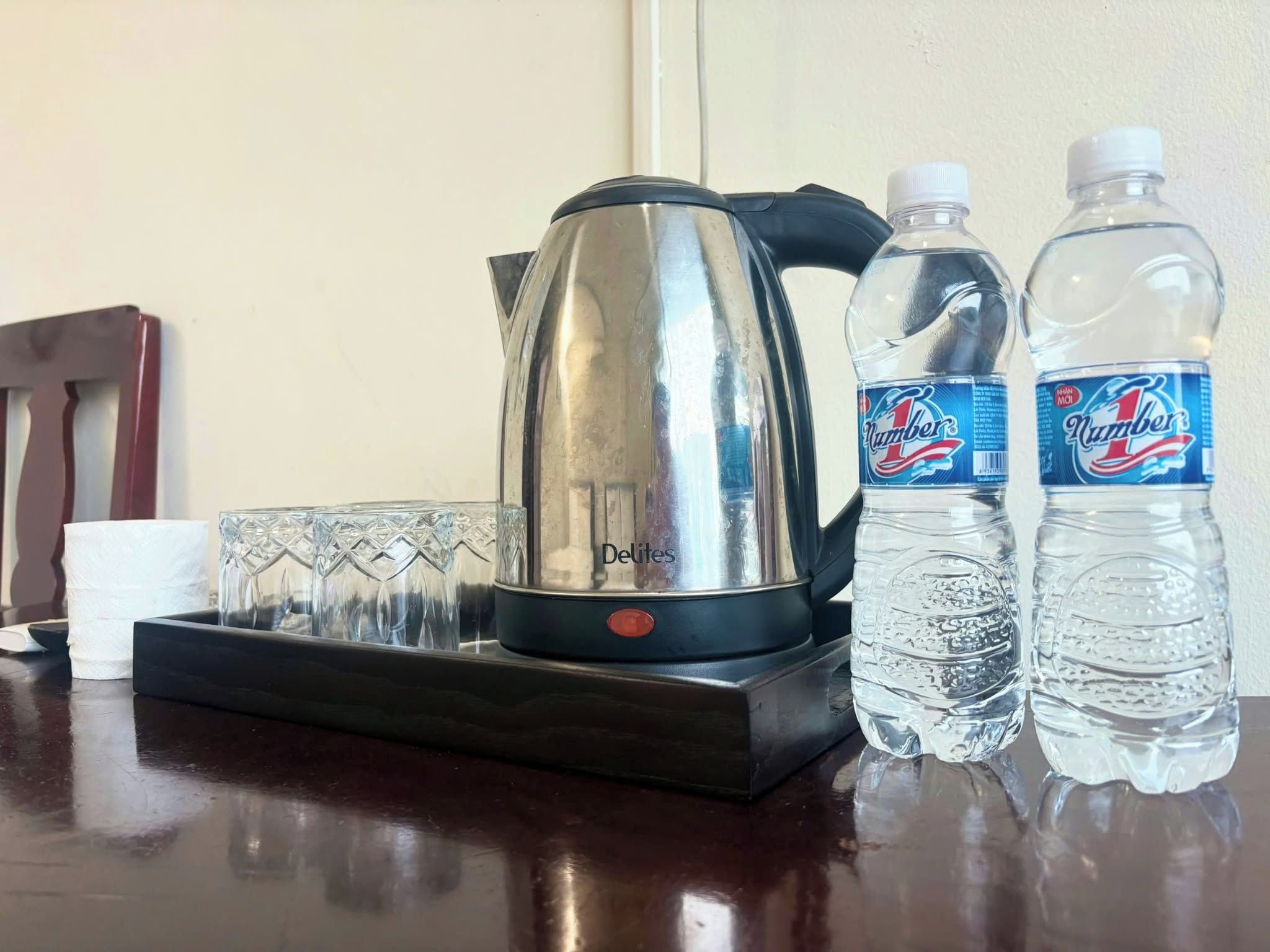 Uma mesa com chaleira, duas garrafas de água, conjunto de copos e rolo de lenços.