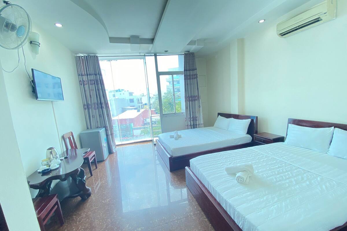A hotel room with two beds, a desk with a chair, a TV, and a large window with curtains.