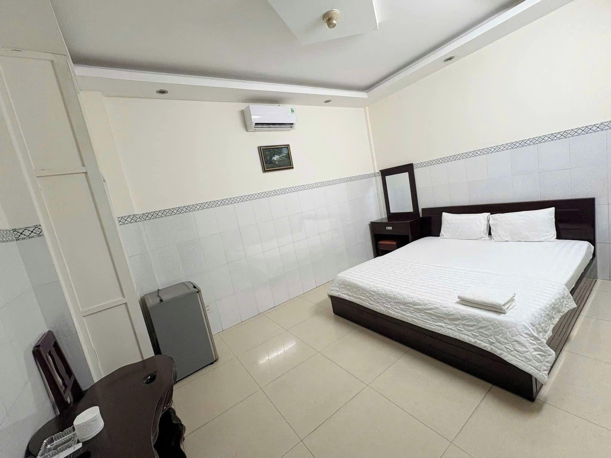 A hotel room with a bed, a desk, a chair, a suitcase, and an air conditioning unit.