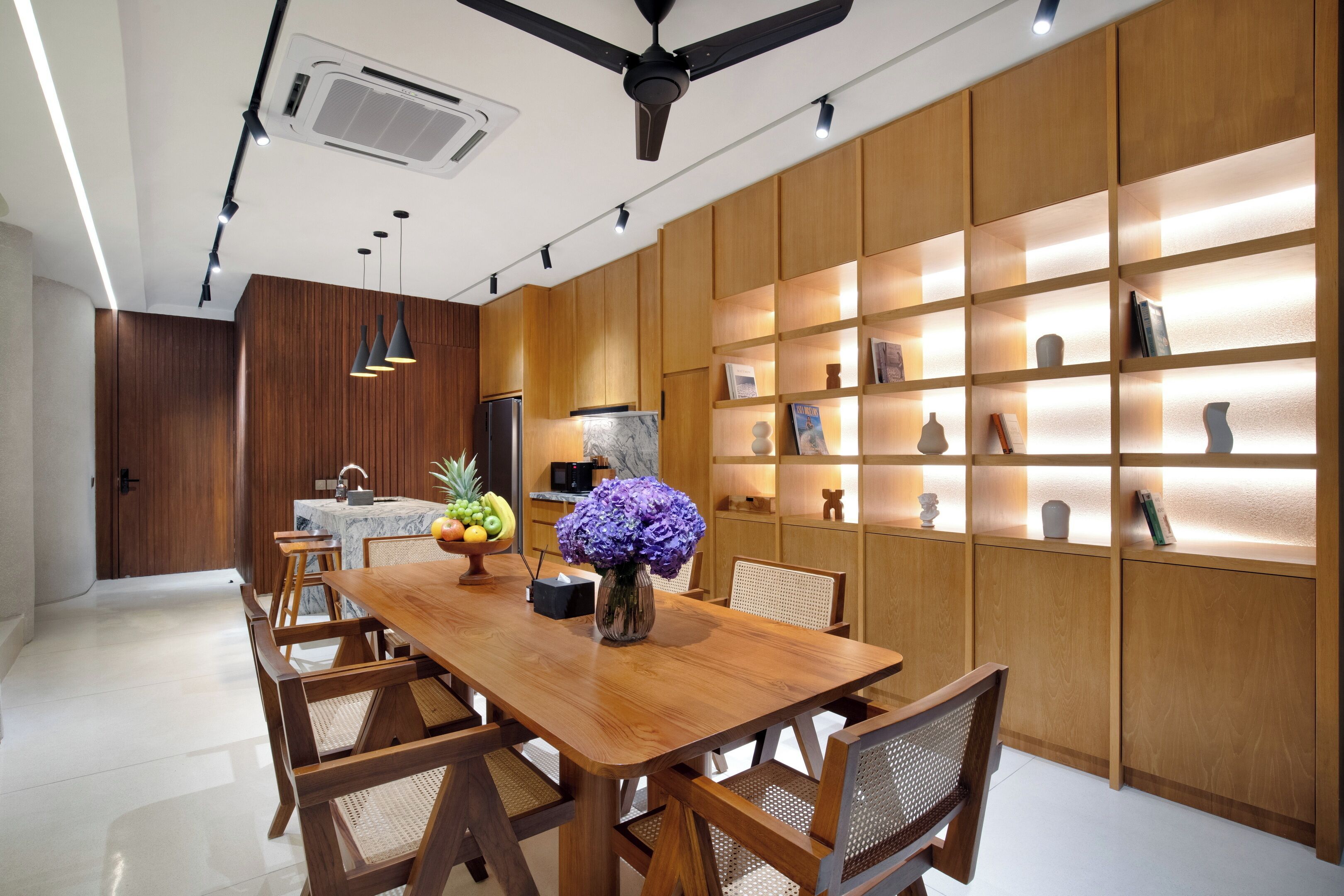 Un espace repas moderne avec une table et des chaises en bois, une cuisine en arrière-plan et des étagères éclairées où sont exposés des objets décoratifs.
