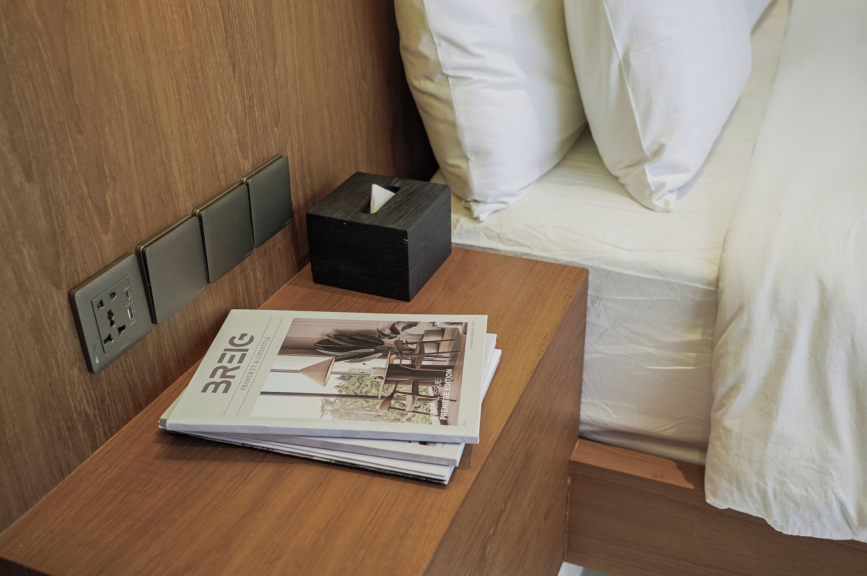 A neatly made wooden bedside table with a tissue box and a stack of magazines.