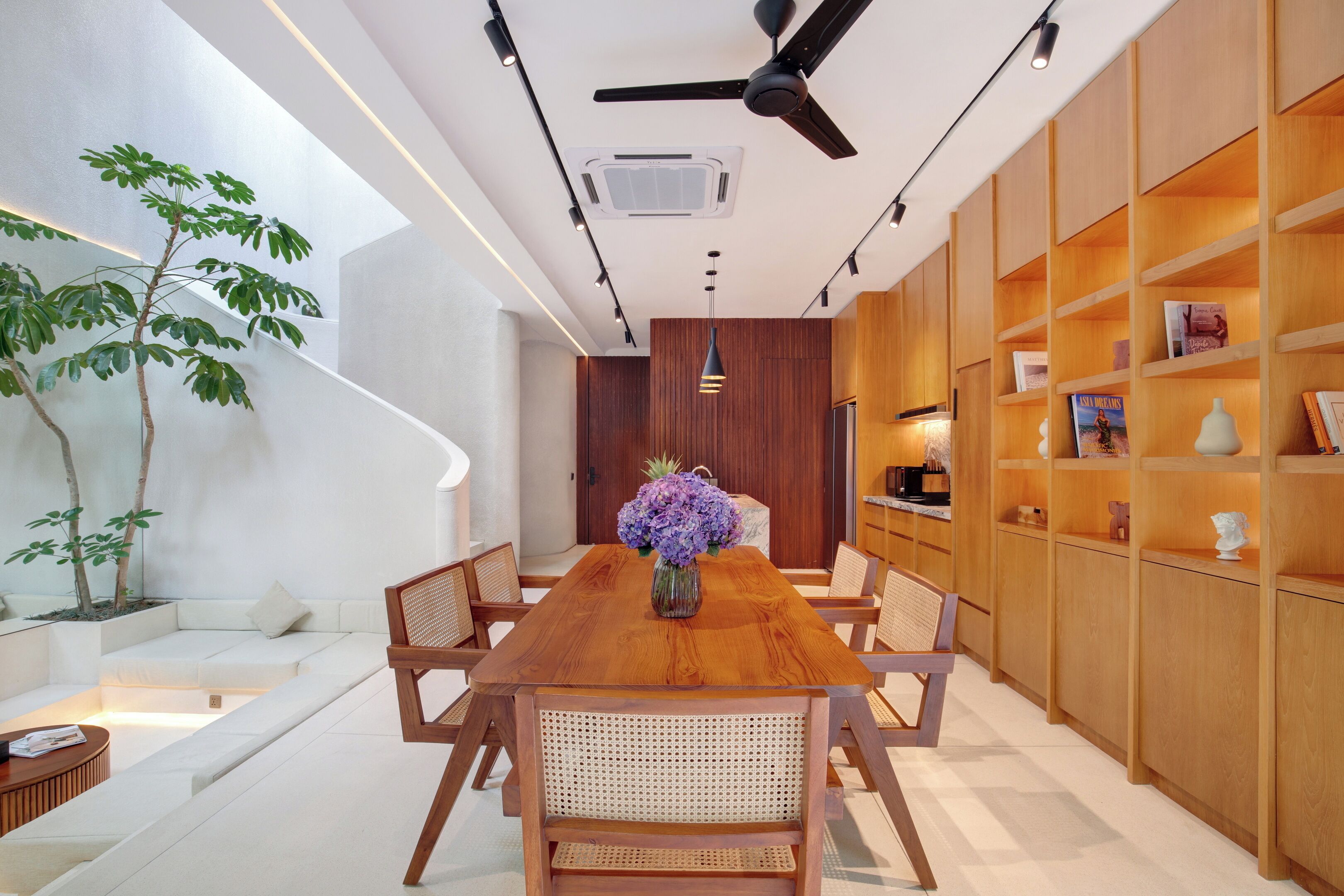 A modern dining area with a wooden table, chairs, and a vase of flowers. There are plants and bookshelves in the background.