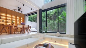 A modern living room with a sofa, a coffee table, and a TV. There is a large window with a view of greenery outside. - Suave Villa Canggu (Canggu)