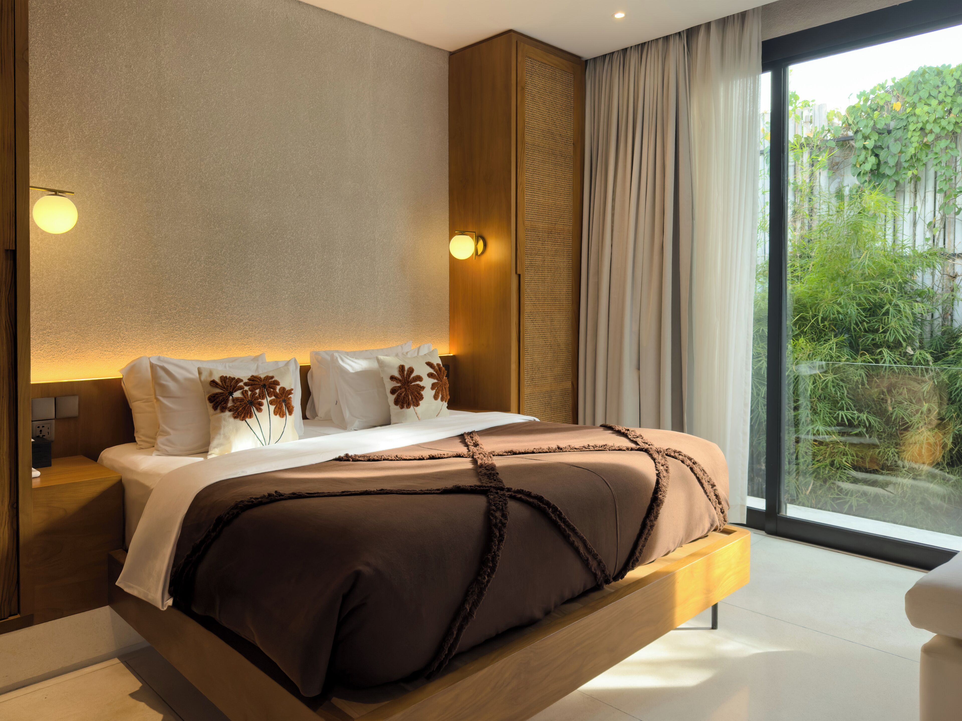 Ein modernes Schlafzimmer mit einem großen Bett, einem Holz-Kopfteil und Blick auf Grün hinter einer Schiebeglastür.