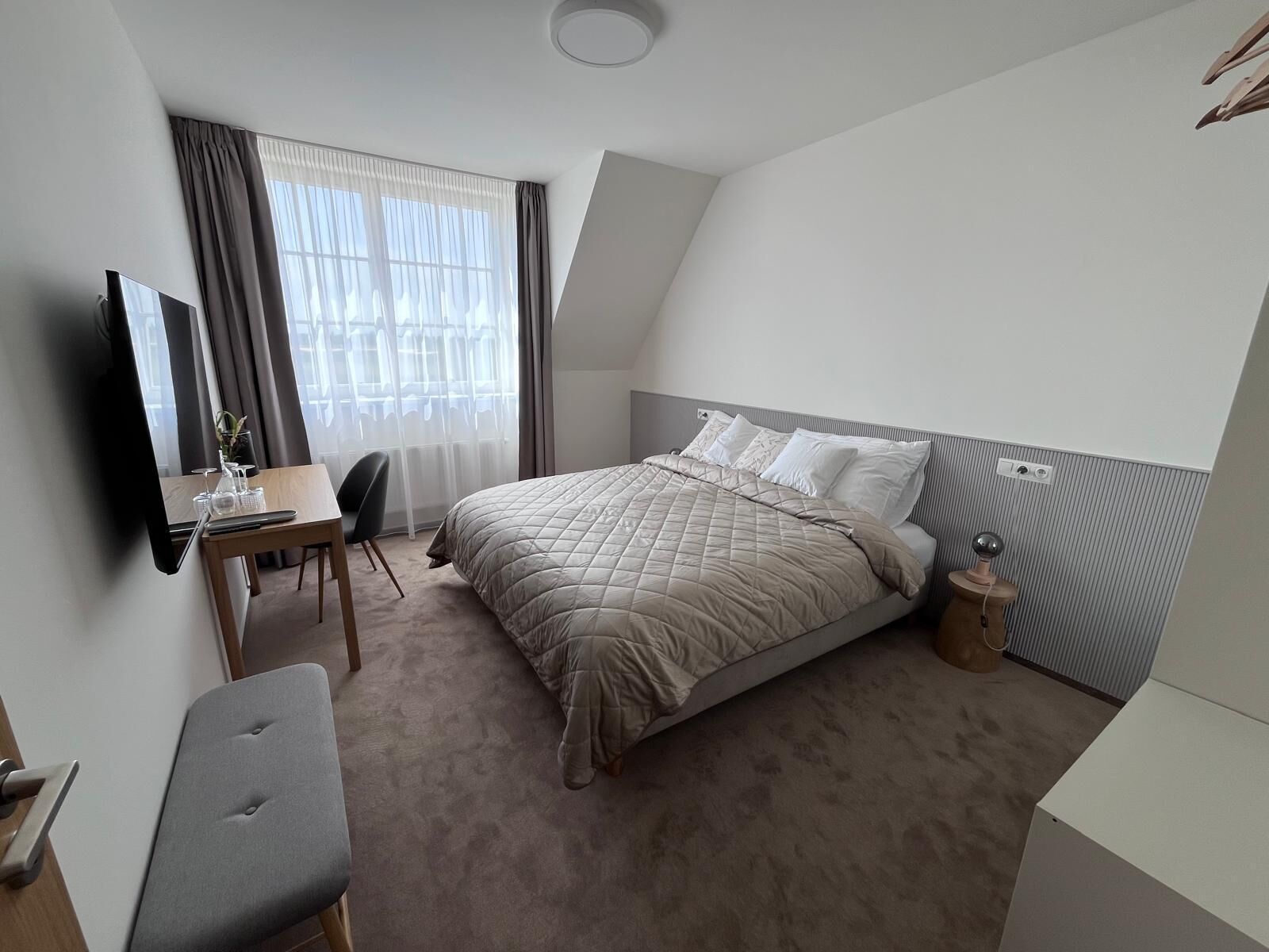 Una habitación de hotel con cama, un escritorio con silla, televisión y ventana con cortinas.