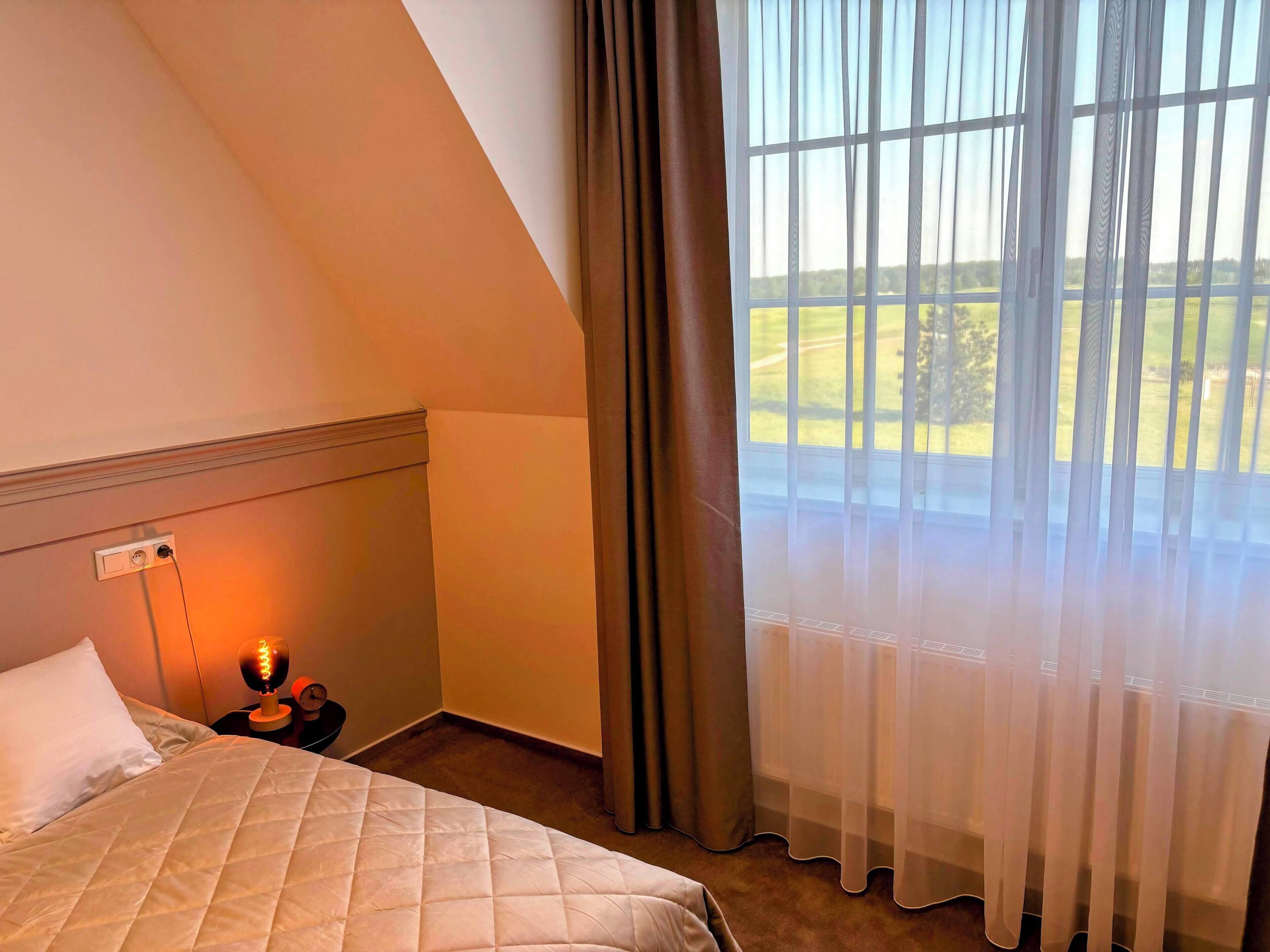 A hotel room with a bed, a nightstand, a lamp, a window with curtains, and a view of a green landscape.