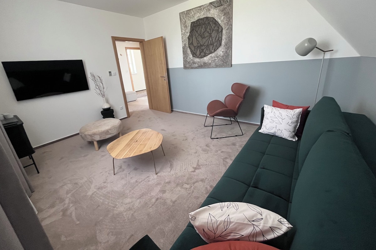 Un salon avec un canapé vert, une table basse en bois, un fauteuil marron et un téléviseur à écran plat fixé au mur.