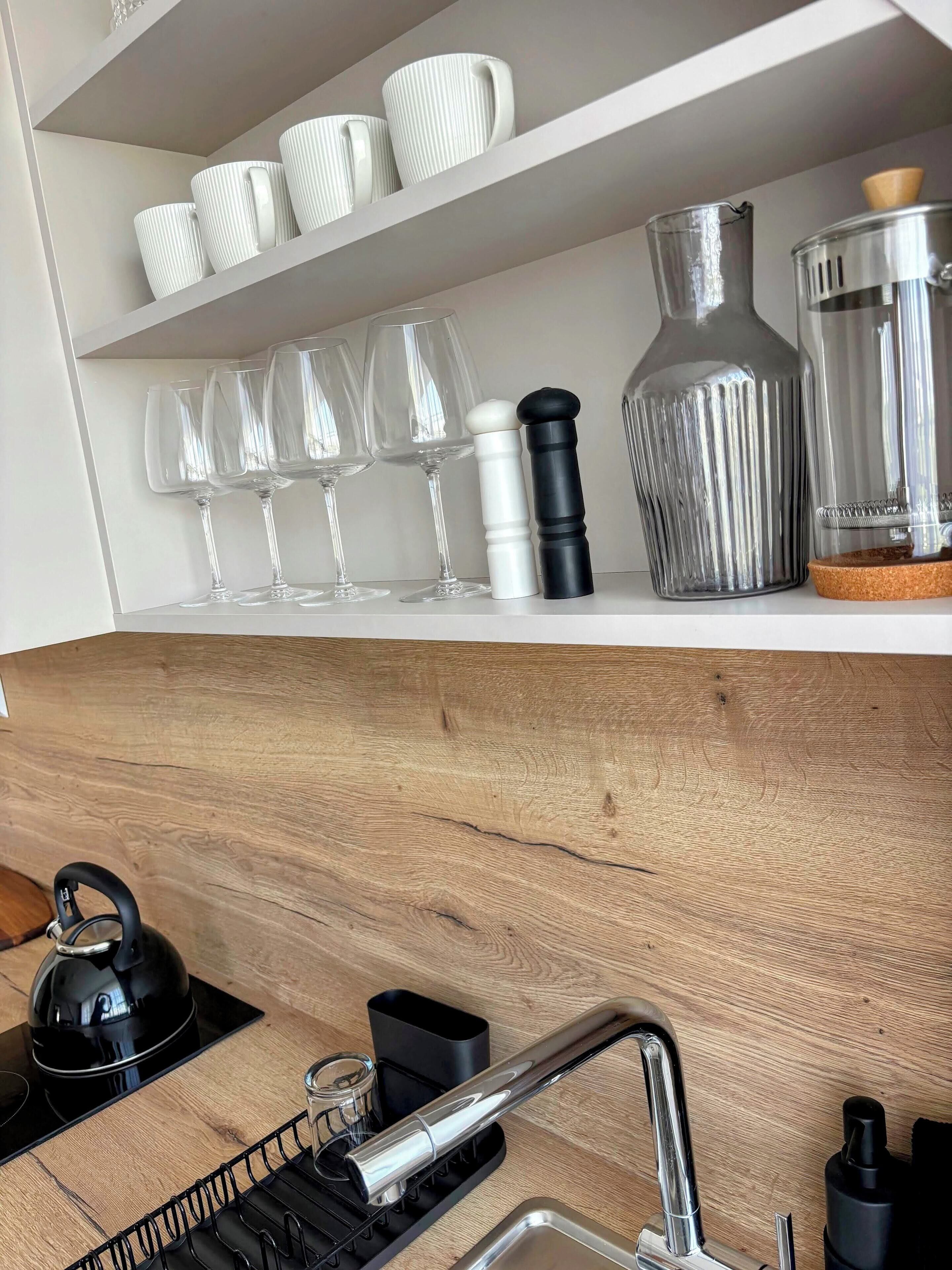 A kitchen with white shelves holding white mugs, clear wine glasses, a black pepper grinder, a glass jar, and a metal container. Below, there's a wooden countertop with a kettle, a coffee maker, a sink, and a black tray.
