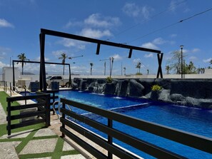 Une piscine entourée d’une clôture en métal noir, avec des palmiers et un ciel d’un bleu limpide.
