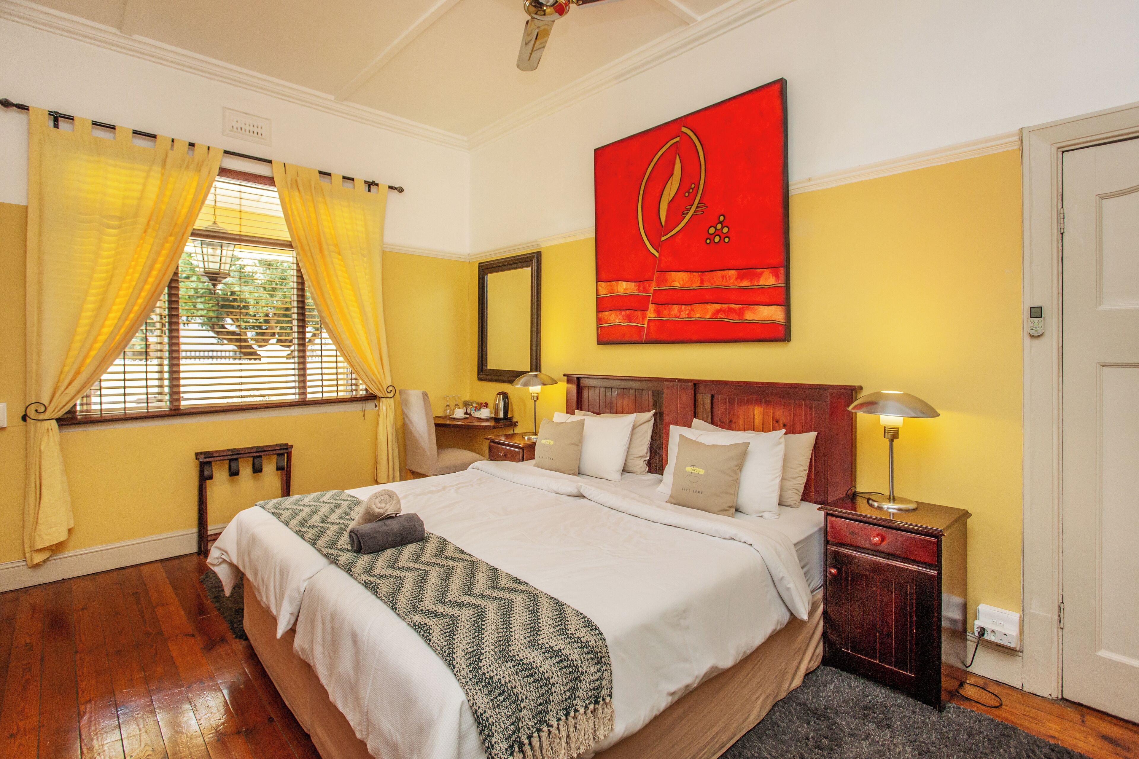 A bedroom with a large bed, wooden headboard, a nightstand with a lamp, a mirror on the wall, and a window with yellow curtains.