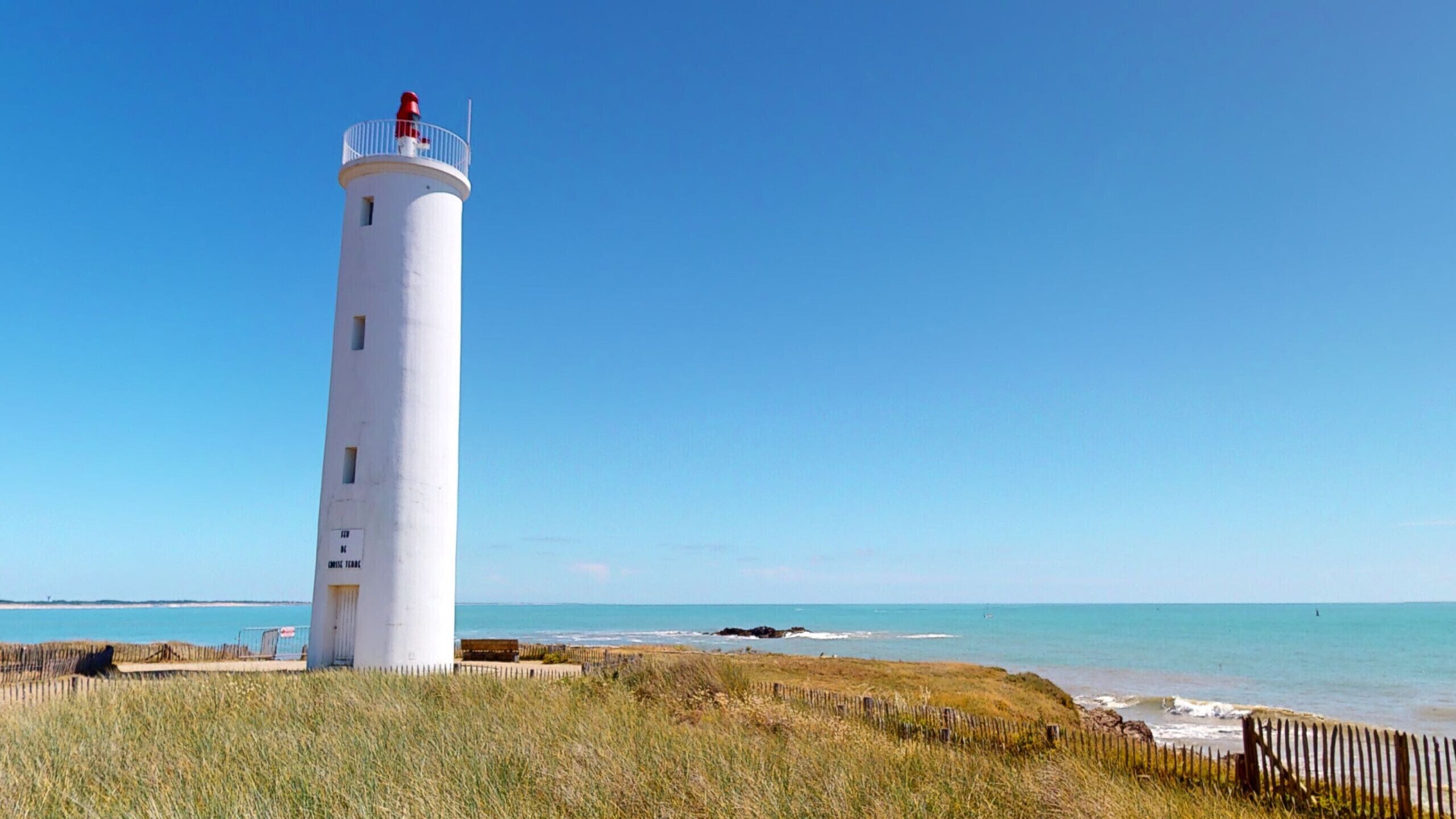 A tall white lighthouse with a red top, situated on a grassy area near a beach with clear blue skies.