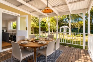 A covered outdoor dining area with a table set for four, surrounded by chairs, and a view of a garden with trees and a gazebo.