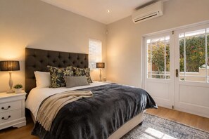 A bedroom with a large bed, two bedside tables with lamps, a window with blinds, and an air conditioning unit.