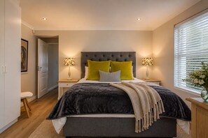 A bedroom with a large bed, two bedside lamps, a window with blinds, and a door leading to another room.
