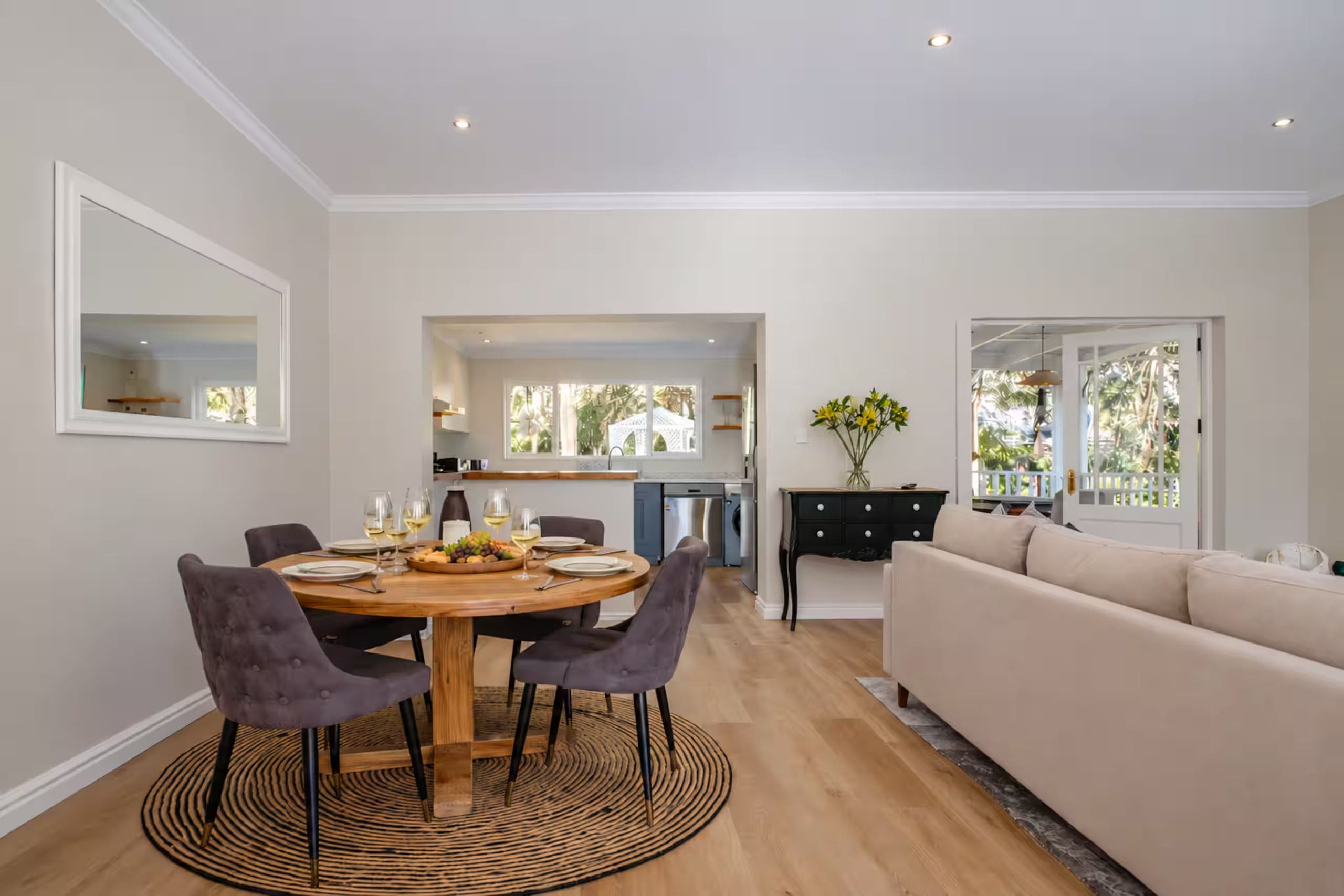 A modern living room with a dining area, a kitchen in the background, and a large window providing natural light.