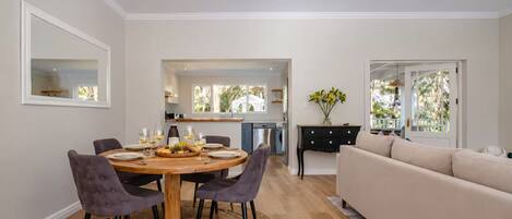A modern living room with a dining area, a kitchen in the background, and a large window providing natural light.