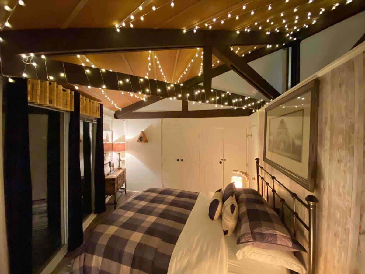A bedroom with a checkered bedspread, a bedside table with a lamp, and string lights on the ceiling.