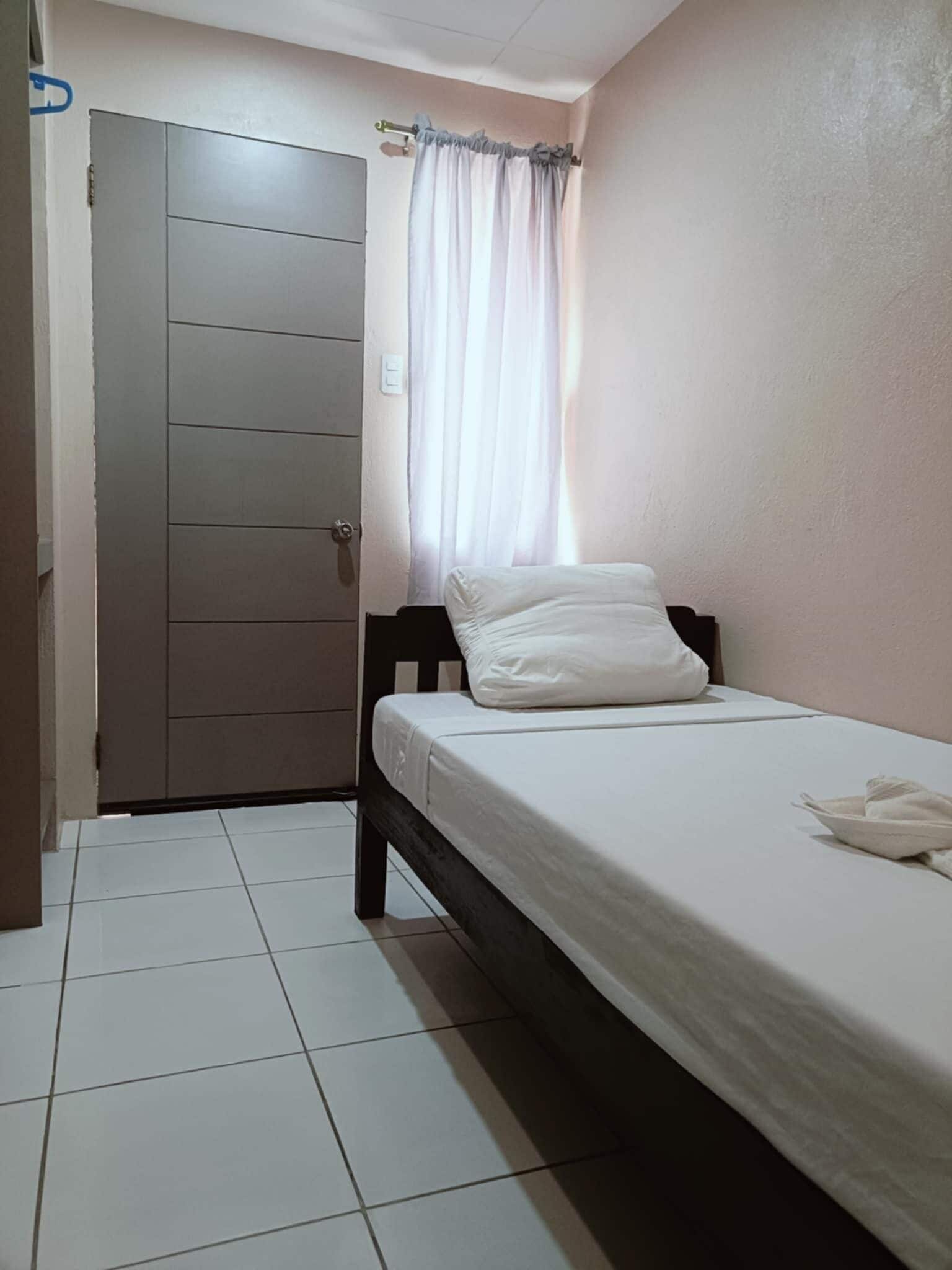 Ein kleines, sauberes Zimmer mit einem Einzelbett, einer Tür und einem mit Vorhängen versehenen Fenster.