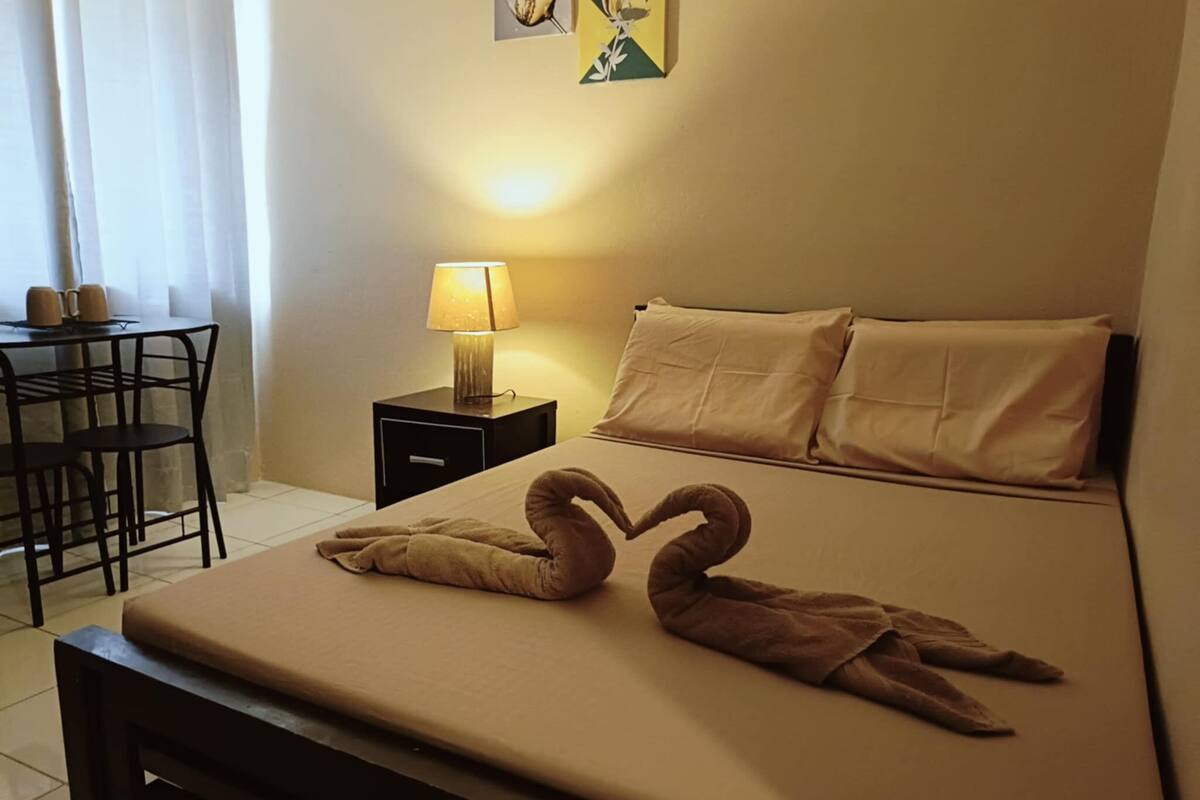 A hotel room with a bed, a nightstand with a lamp, a chair, and a table with cups.
