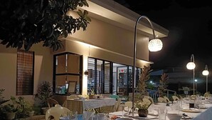 Un área de comedor al aire libre con mesas dispuestas para una comida, rodeada de plantas en macetas y un edificio con puerta de vidrio al fondo.