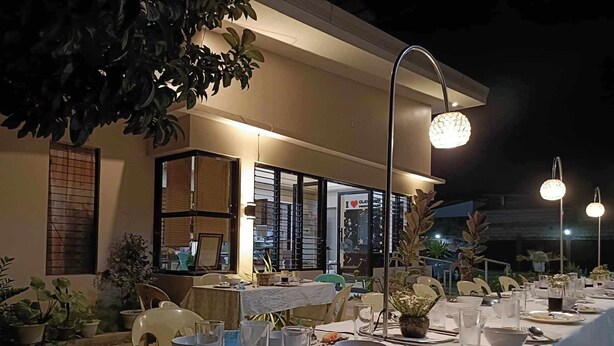 Un área de comedor al aire libre con mesas dispuestas para una comida, rodeada de plantas en macetas y un edificio con puerta de vidrio al fondo.