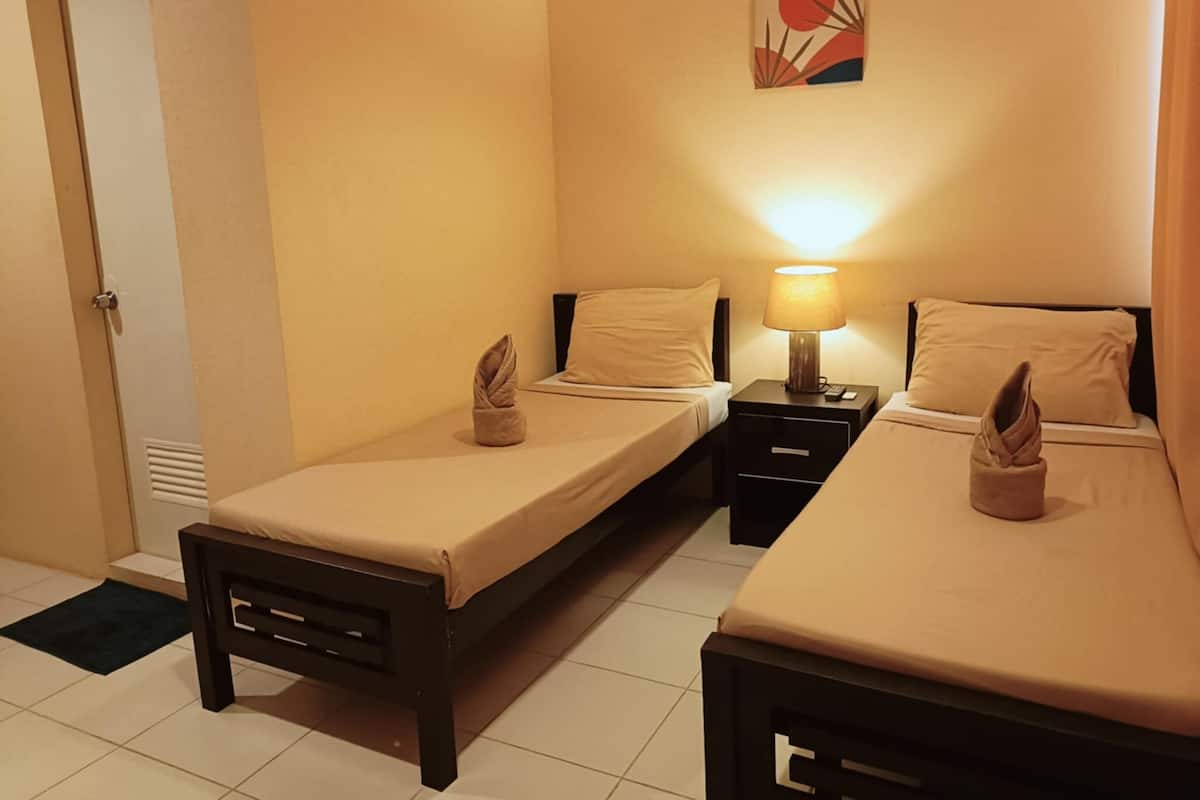 A hotel room with two single beds, a bedside table with a lamp, a door, and a wall-mounted artwork.