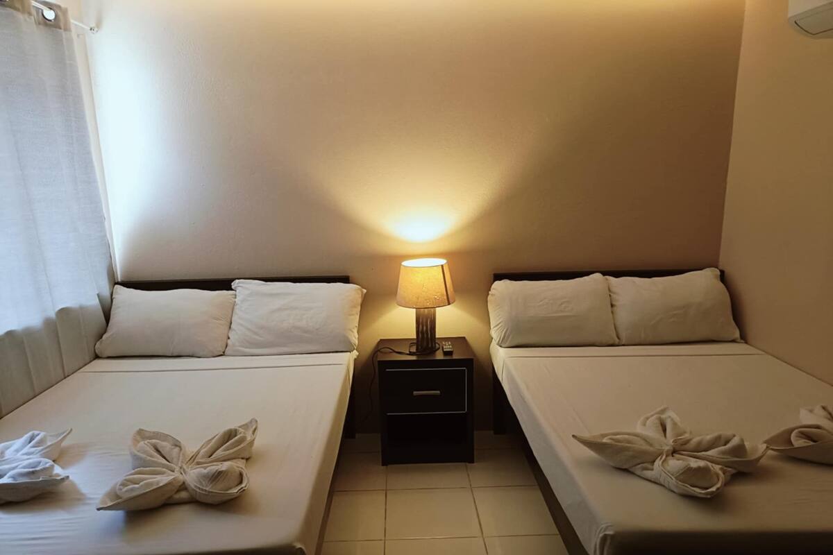 A hotel room with two beds, each with white linens and towels arranged in a bow shape. There is a nightstand with a lamp between the beds.