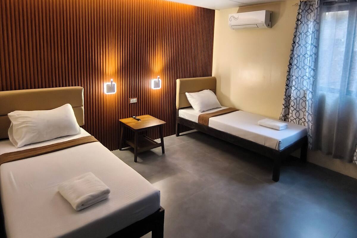 A hotel room with two beds, a small table, a wall-mounted air conditioner, and a window with curtains.