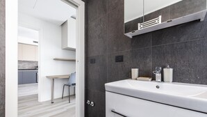 A modern bathroom with a white sink, dark tiled walls, and a visible door leading to a kitchen area. - East Uptown P Pasolini 74 Cascina Merlata (Milan)