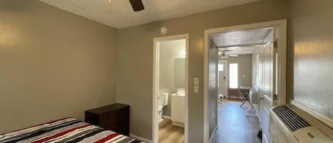 Quarto com cama, ventilador de teto e corredor que dá para um banheiro.