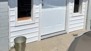 Une porte blanche avec deux fenêtres de chaque côté, un seau en métal posé sur le sol et une voiture garée à proximité.