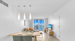 A modern living room with a dining area, a TV, and a large window with a city view. - Infinite View, Amazing Apt, Spectacular Amenities (Hallandale Beach)