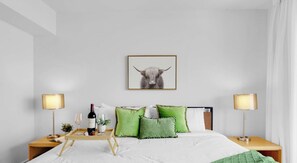 Un dormitorio ordenado con cama, mesitas de noche, una lámpara, una bandeja con copas de vino y un cuadro en la pared.