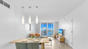A modern living room with a dining area, a TV, and a large window with a city view. - Spectacular Apt, Infinity View, Amazing Amenities (Hallandale Beach)