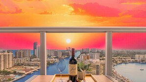 A bottle of wine on a tray with a glass, set against a sunset view from a high-rise window overlooking a cityscape and water. - Spectacular Apt, Infinity View, Amazing Amenities (Hallandale Beach)