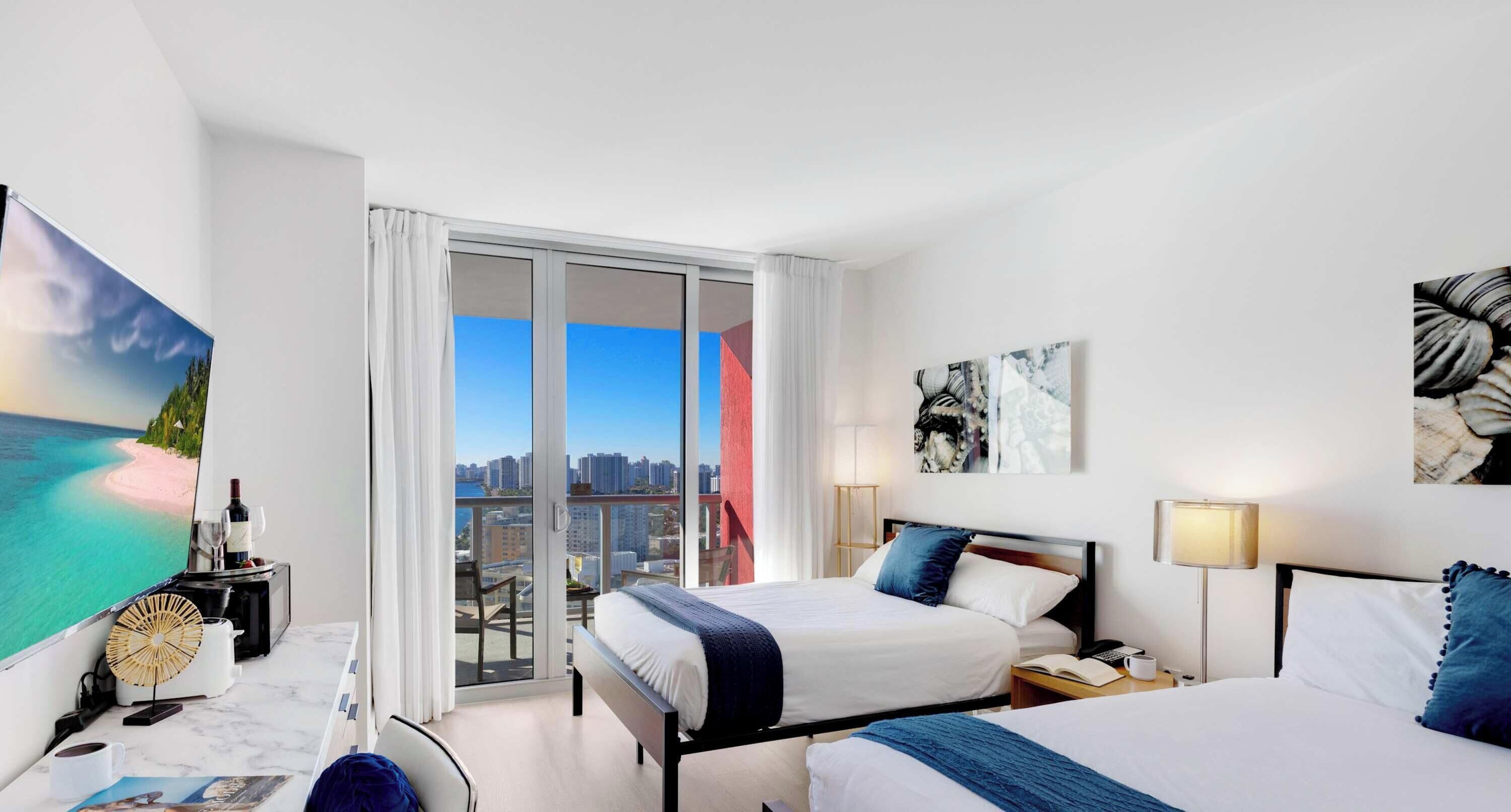 A hotel room with a bed, bedside table, lamp, and a view of the city through large windows.