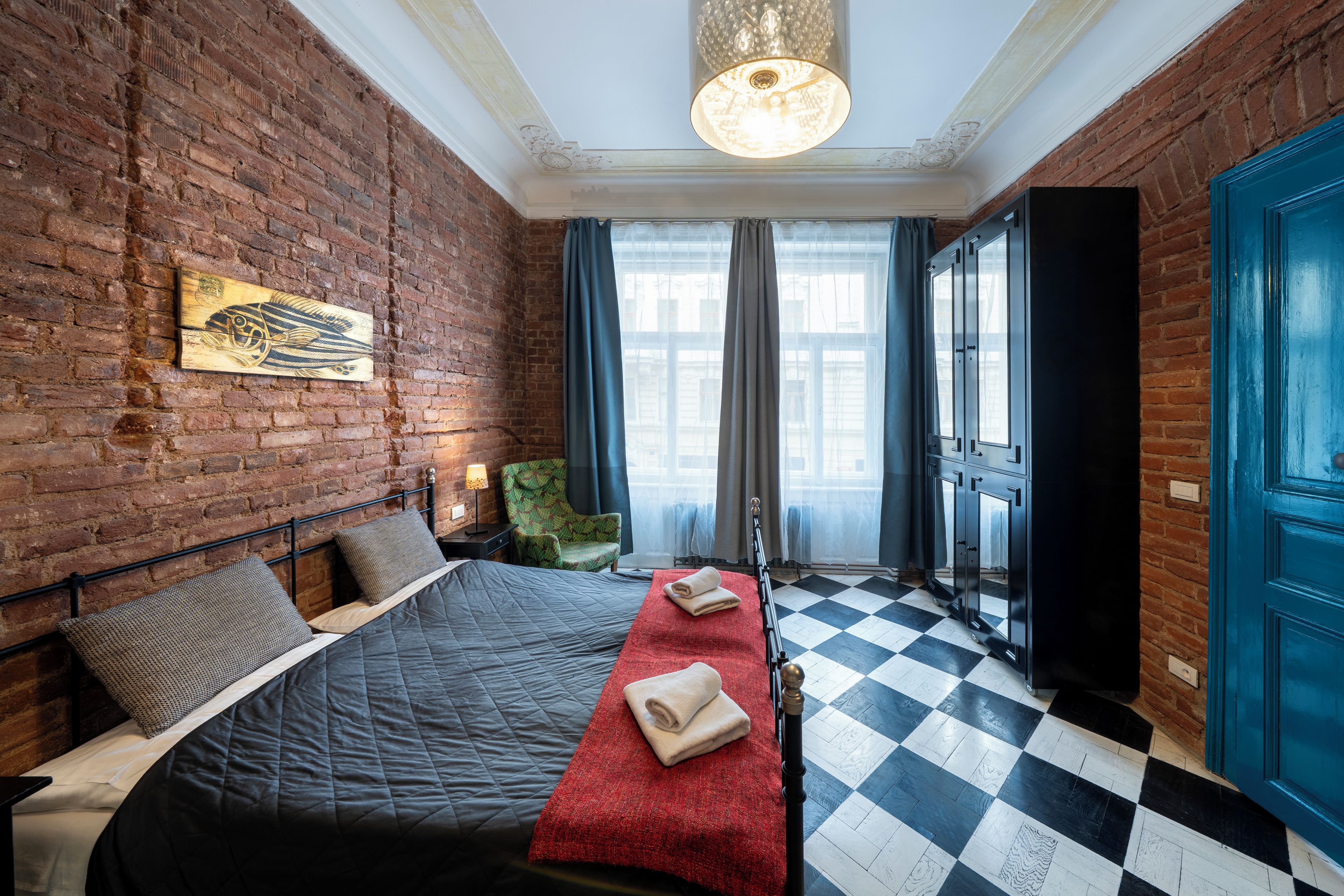 A hotel room with a brick wall, a bed with a red runner, a checkered floor, and a blue door.