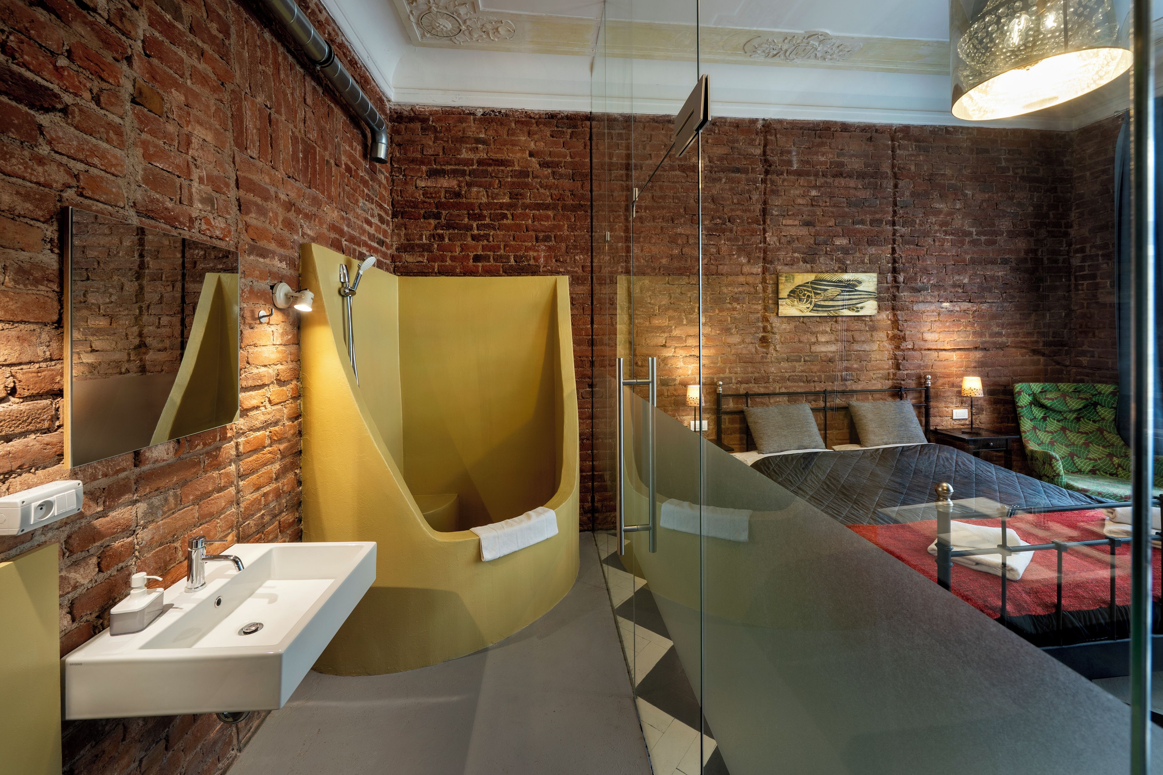 Een moderne badkamer met een bakstenen muur, een geel bad, een douchecabine van glas en een bed met een bedsprei met een groen patroon.