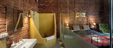 A modern bathroom with a brick wall, a yellow bathtub, a glass shower enclosure, and a bed with a green patterned bedspread.