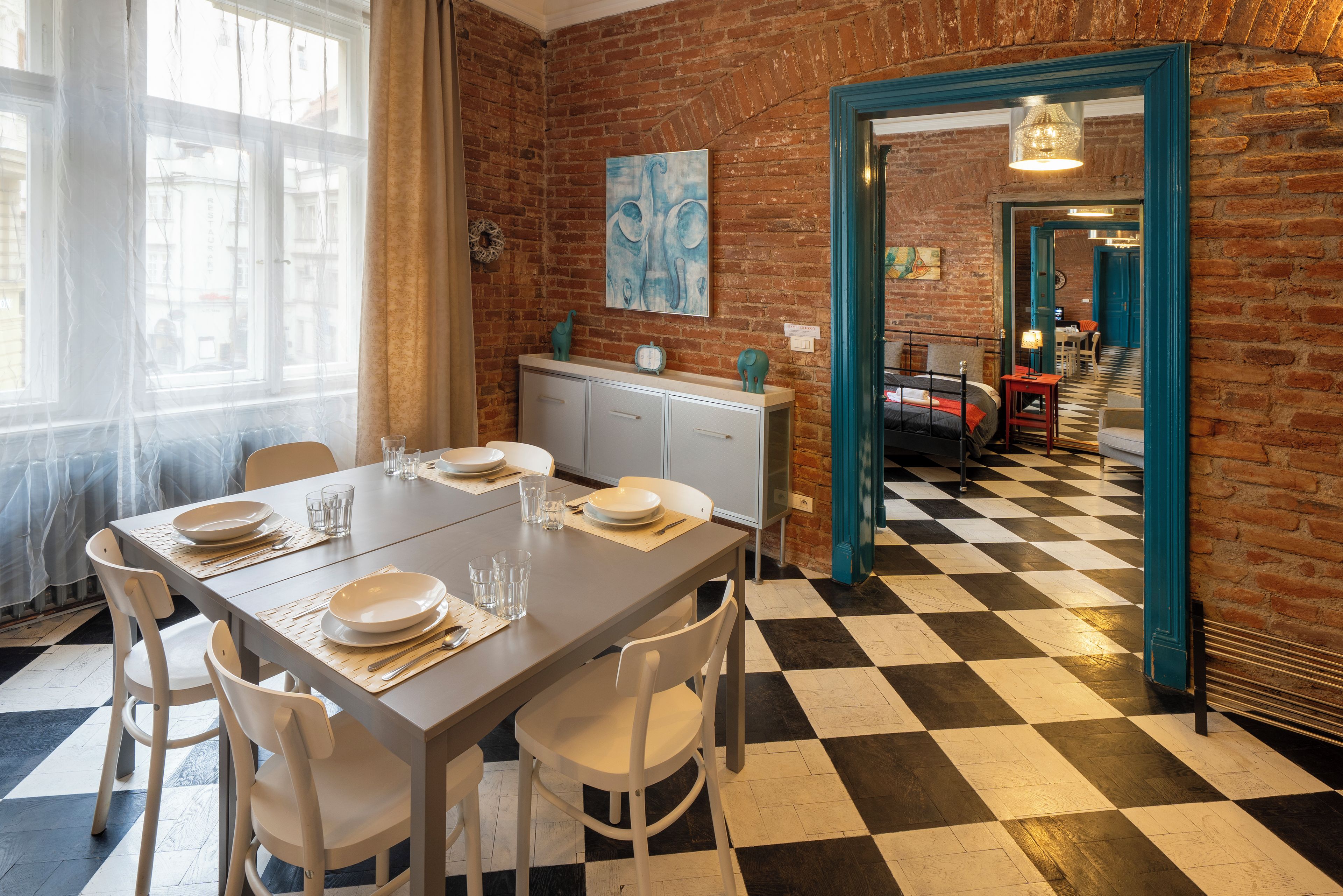 Un comedor con piso a cuadros, sillas blancas, una mesa lista para cuatro personas y una pared de ladrillo con un cuadro.