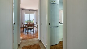 Un couloir mène à une salle à manger avec une table et des chaises, une cuisine équipée d’un réfrigérateur et une fenêtre avec des rideaux.