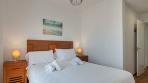 Un lit bien fait, avec du linge de lit et des serviettes blanches, une tête de lit en bois, deux lampes de chevet et un tableau représentant une scène de plage accroché au mur.