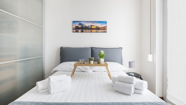A modern hotel room with a bed, a bedside table, a nightstand, a window, and a painting on the wall. - Easylife - Modern apartment in Bocconi (Milano)
